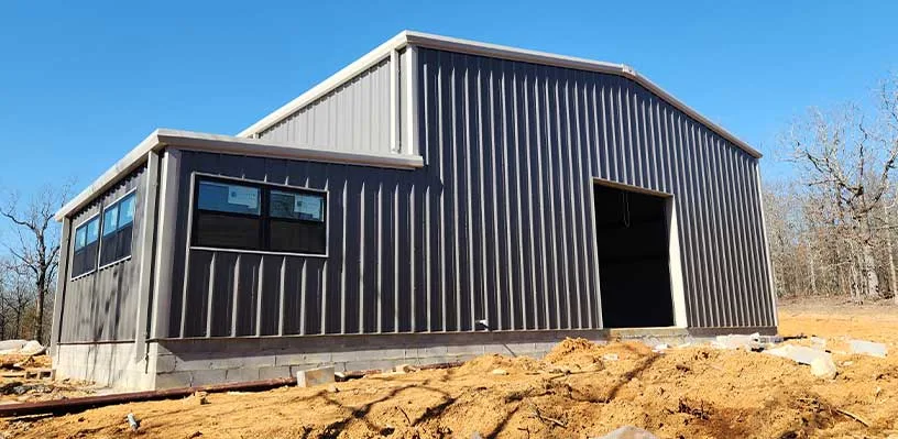 Gray metal building with sloped roof on dirt lot, features windows and open entrance. Sparse trees.