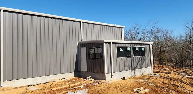 Gray metal building with door and windows in wooded area, dirt ground in the foreground.
