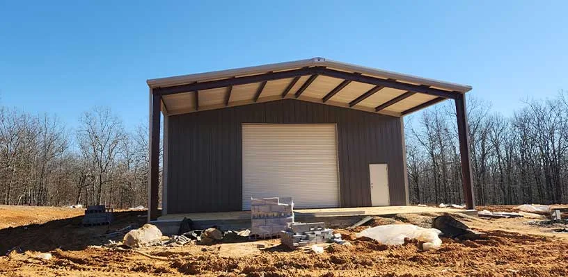 Metal building with overhead door, side door, construction materials, bare trees, clear sky.