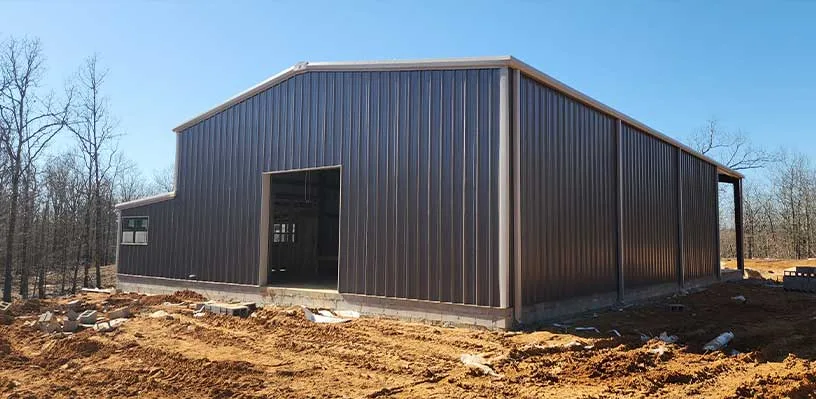Metal building under construction on dirt site with leafless trees in the background.