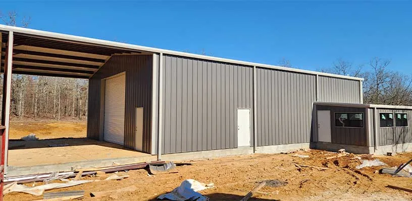 Large metal building with high roof, roller door, windows, surrounded by leafless trees under blue sky.