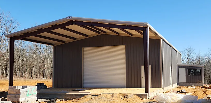Steel structure with gabled roof, roll-up door, construction site in clearing, sparse trees.